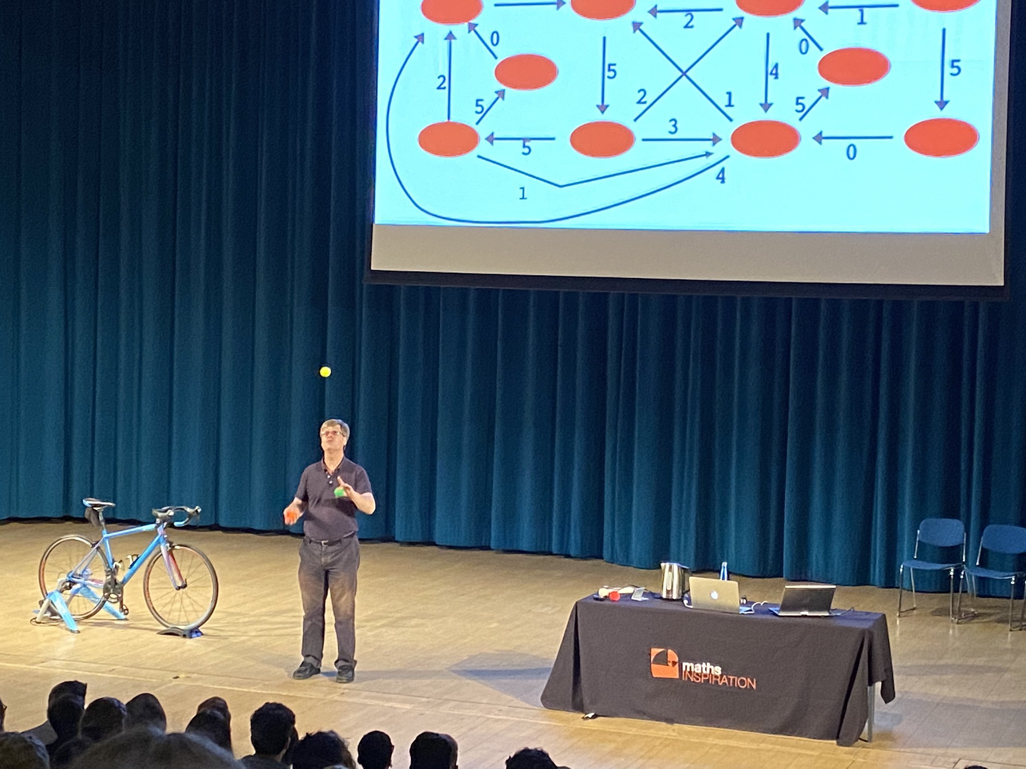 Photo of a theatre stage, with a blue curtain behind, a slide with a complicated maths diagram on screen, and a presenter who is a tall white red-haired man standing on stage juggling next to a table covered in objects