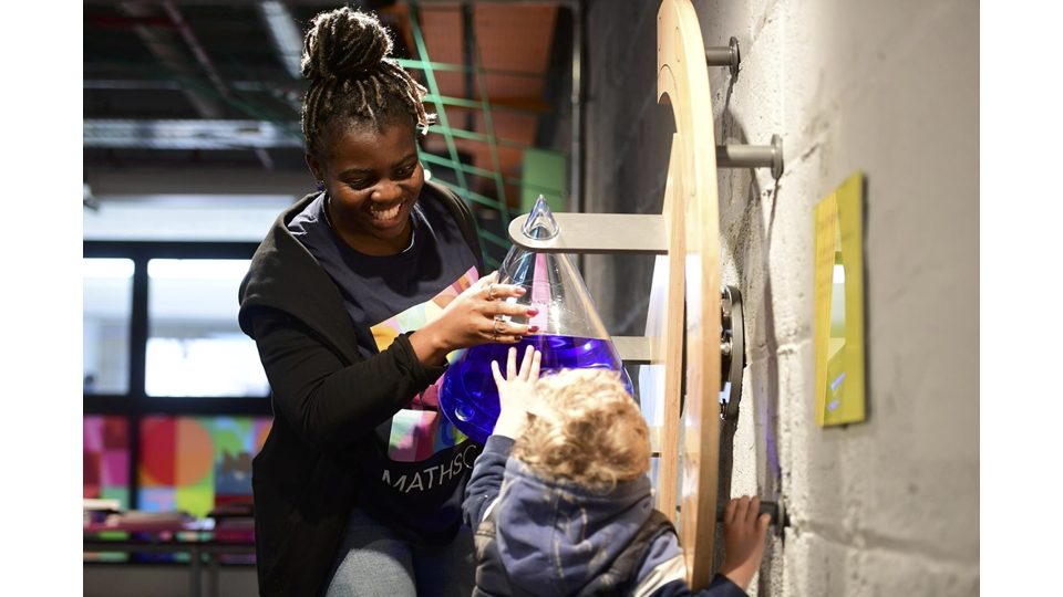 A photograph of a small visitor and a member of the MathsCity explainer team, looking at a cone filled with liquid to demonstrate conic sections