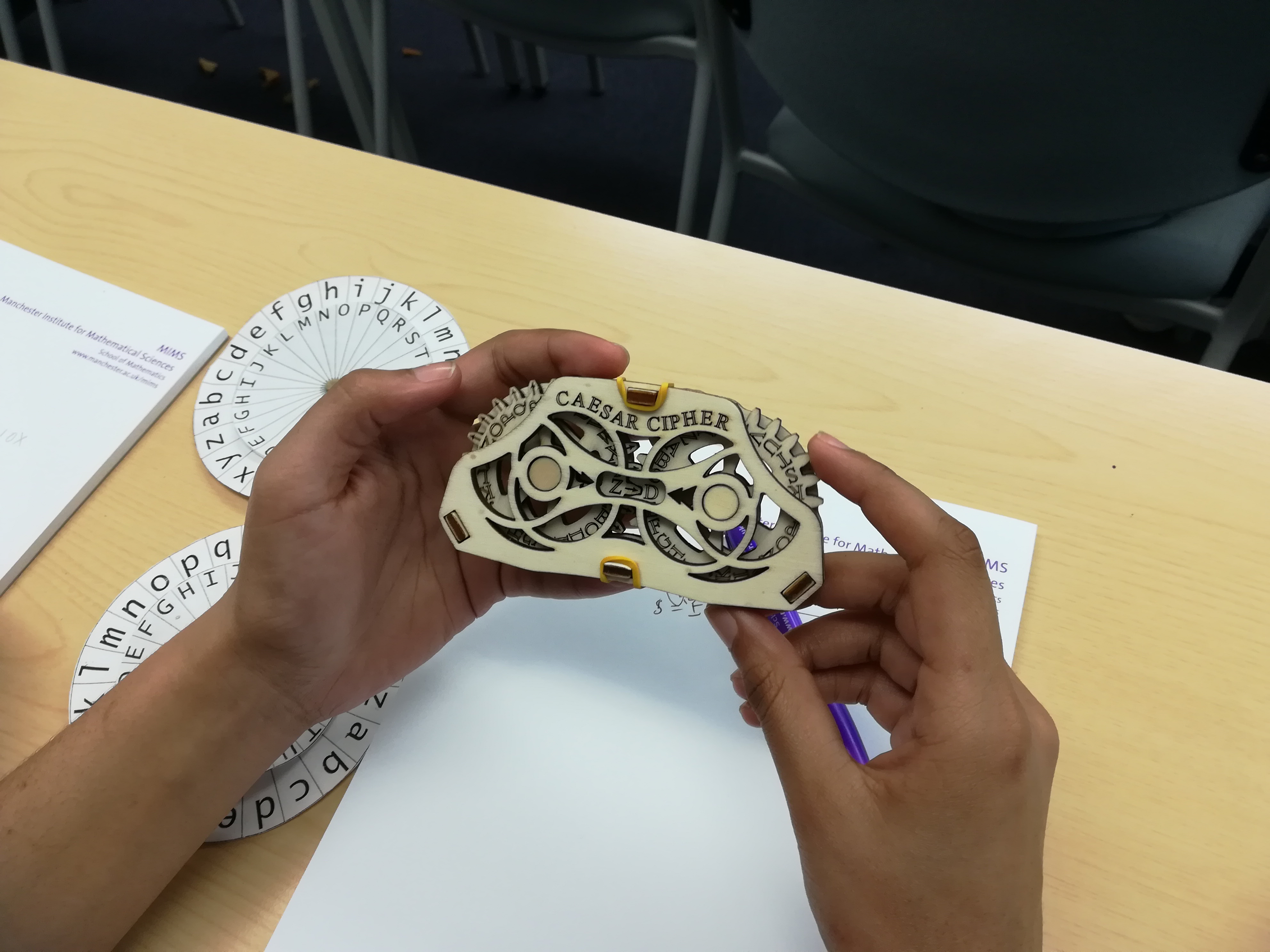 A photograph of a student’s hands holding a wooden Caesar cipher solving tool, with some decoding wheels on the table behind