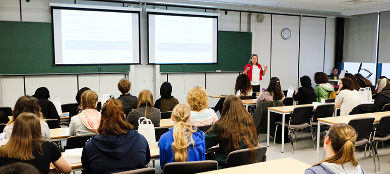 A mathematician (Professor Louise Walker) standing in front of a room of young students, with slides on the projector and talking animatedly